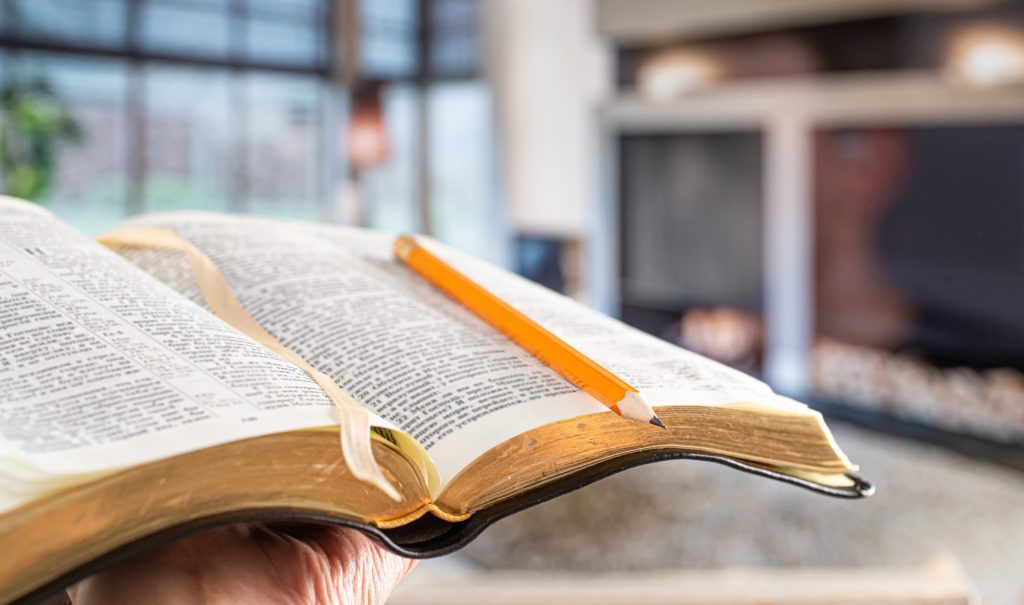 A man holds a Bible with a pencil, against the background of the living room.
